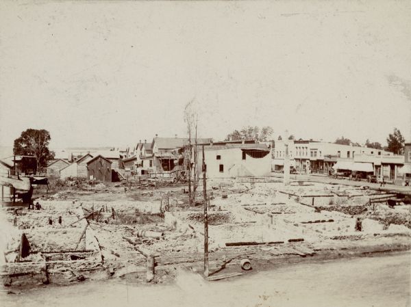Elevated view looking west over the open basements of commercial buildings which were destroyed by a recent fire. Main Street is in the foreground. Horse-drawn wagons are parked along the north side of Nebraska (now West Montello) Street, on the right, in front of commercial buildings spared from fire. Pedstrians are walking on the sidewalk. The exposed side wall of the building facing Nebraska street shows fire damage and partial collapse. Buffalo Lake is seen in the background on the left.