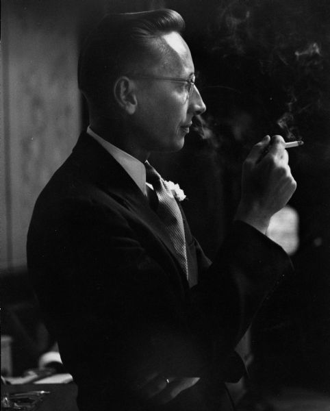 Oliver Justman, looking suave, calmly smokes a cigarette as he watches his bride Dorothy being photographed.