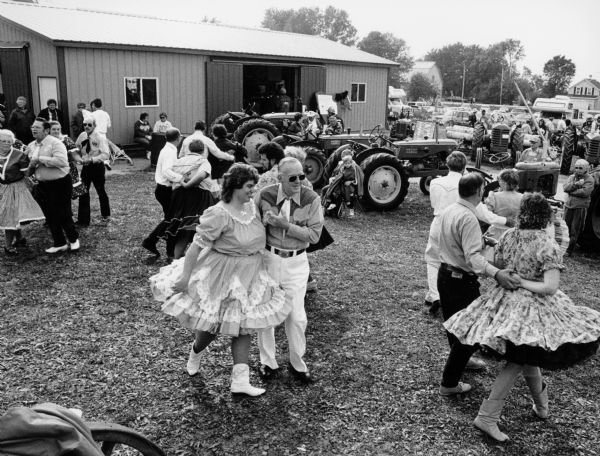 Square dancers in farmyard at Calumetville. Barn and tractors can be seen in the background.