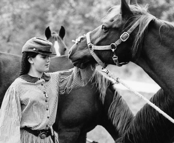 A young woman civil war re-enactor pets a horse. More horses can be seen in background.