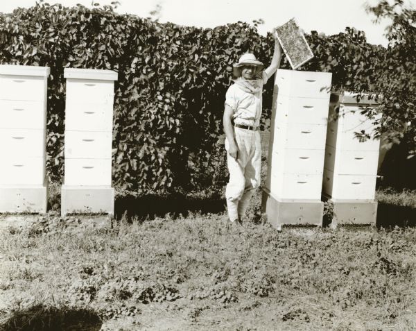 A young man wearing a veiled hat stands next to hives while holding up a frame with bees on it. He holds a hive tool in his right hand.