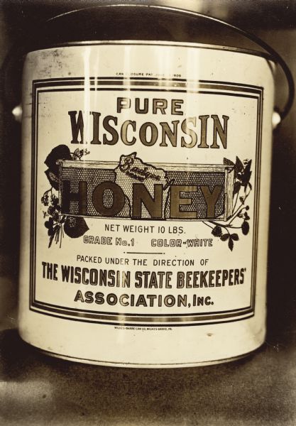Ten pound can of honey. The label reads: "Pure Wisconsin Honey, Badger Brand, packed under the direction of the Wisconsin State Beekeepers' Association, Inc." The word "Honey" is over an illustration of a frame with honeycomb, the shape of the state of Wisconsin also as a honeycomb, and bees flying near clover flowers on the right and left. Writing on back of print: "Lithographed Part of Wis. Assn."