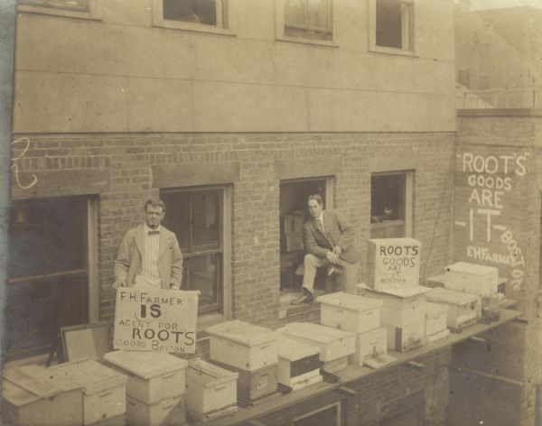 Two men are posing standing on a balcony on the side of a brick building with several beehives. The man on the right is in front of an open window and is holding a hive smoker. There are hand-painted advertisements for Root's Goods — selling beekeeping supplies, local dealer F.H. Farmer.