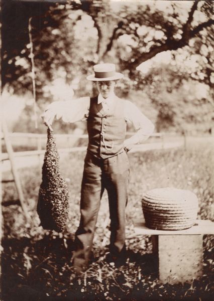 Young man standing next to a bee skep while holding a swarm of bees. German writing on back: "Mein John Felix."