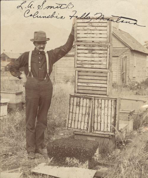 Beekeeper L.A. Smith standing next to hive boxes stacked on top of each other, exposing the frames. There is a swarm of bees on a hive box on the ground.