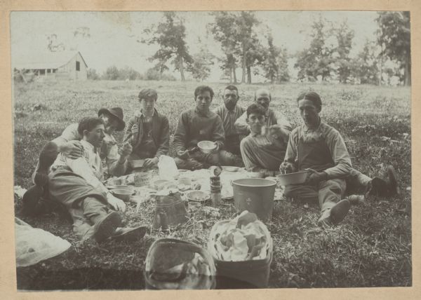 A group of young men seated on the ground, having a picnic. A can of honey, salmon, and bread are visible on the ground. Two of the men have metal dots on the bottom of their boots – might be significant in beekeeping work.
