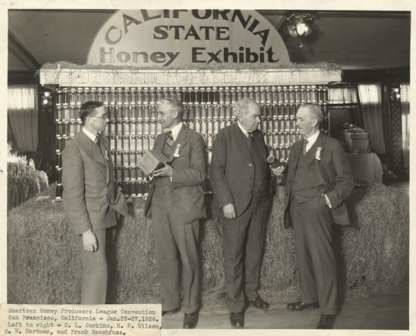 Group portrait of four men: C.L. Corkins, H.F. Wilson, C.W. Hartman, and Frank Rauchfuss standing in front of a display of honey in jars, and boxes of honeycomb. A sign above the exhibit says: "California State Honey Exhibit." Taken at the American Honey Producers League Convention.