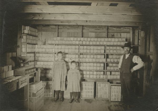 G.W. Estes and two young girls, standing in a storeroom. Hive boxes with frames are on the floor. Along the walls are shelves stacked with square boxes of comb honey. A work table on the left has a scale.