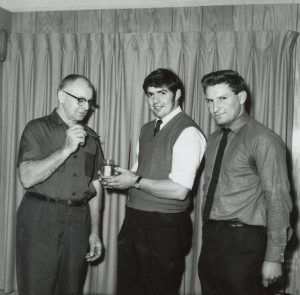 Three men standing in front of a window curtain sampling honey.