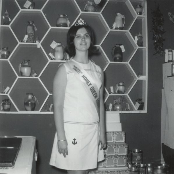 Manitowoc County Honey Queen, Lois Lensmeyer of Whitelaw, Wisconsin. She wears a white dress and is standing and smiling with her Honey Queen sash and crown, working in the Wisconsin Honey Producers Booth at the Wisconsin State Fair. Ceramic jars of honey are displayed behind Lois on honeycomb styled shelves, and boxes of comb honey are displayed below.