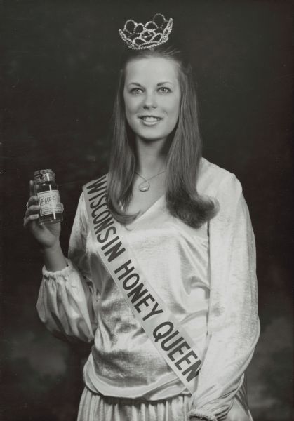 Studio portrait of the Wisconsin Honey Queen holding a jar of honey from Johnson Creek, Wisconsin. She is wearing a tiara and a sash reading "Wisconsin Honey Queen."