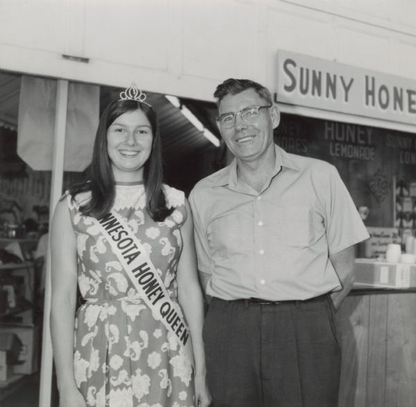 August Laechelt (La Valle, Wisconsin) and the Minnesota State Honey Queen at the Minnesota State Fair. The Honey Queen is wearing a sash and tiara. Behind them is a honey display with a sign reading "Sunny Honey."