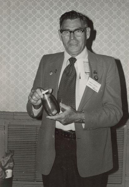 Beekeeper August Laechelt, smiling and holding a jar of honey in his hands. His nametag reads "American Beekeeping Federation; January 18-22, 1976; August Laechelt; LaValle, Wis.; Century Club." A newspaper is in his breast pocket and he wears a variety of pins on his lapel.