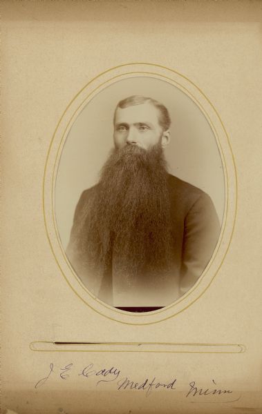Waist-up oval portrait of J.E. Cady, of Medford, Minnesota. He is wearing a suit coat, and has a long beard and a moustache.