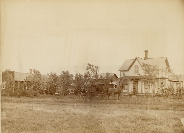 View across field of the house and apiary of J.E. Cady. There is a man in a horse and carriage in front of the house. The carriage has a sign on the side that reads: "Honey." A man is standing in the yard among the beehives. On the far left a building with a false front also has a sign that reads: "Honey."