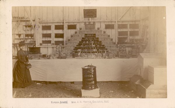 Honey display by the Eureka Apiary. A sign in front of the display on the table says "Hands Off." Mrs. J.N. Heater stands on the left. In the foreground center is Novice's Honey Extractor. Printed on bottom: "Eureka Apiary, Mrs. J.N. Heater, Columbus, Nebraska."