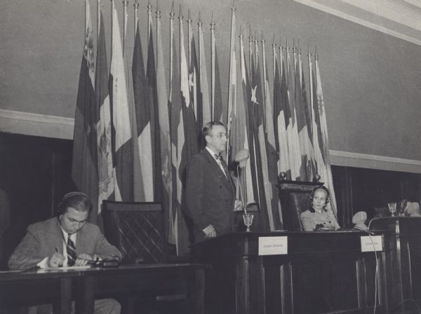 Arthur Altmeyer speaking at the Inter-American Conference on Social Security, which took place in Buenes Aires. Eva Peron is seated to his left, wearing headphones.