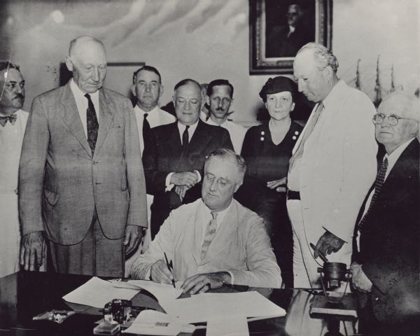 President Roosevelt signs the Social Security Act. Surrounding him are: Robert L. Doughton, Senator Barkley, Senator Wagner, Congressman John Dingell, Secretary of Labor Frances Perkins, Senator Pat Harrison and Representative Lewis of Maryland. On the far left is an unidentified man.