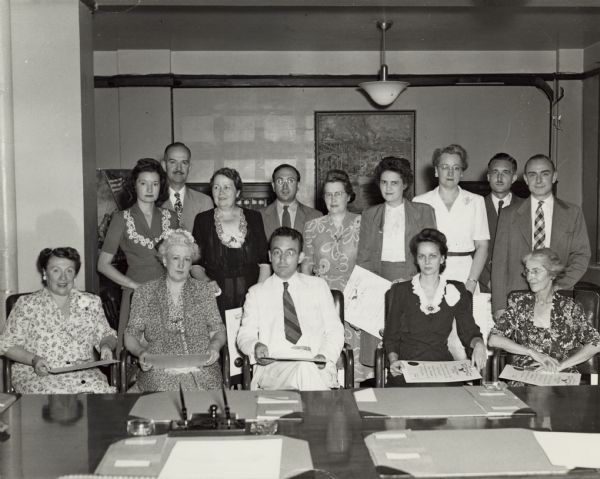 A large group of federal employees celebrating the 10th anniversary of the signing of the Social Security Act.<p>Left to right standing: Evelyn S.  Selecman, Budget Section, Accounting and Budget Division, Bureau of Accounts and Audits; Merrill G. Murray, Bureau of Old-Age and Survivors Insurance, Baltimore, Md.; Naomi L. D'Orsey, Stenographic Section, Personnel and Business Mgm. Services, Office of Exec. Dir.; Wilbur J. Cohen, Bureau of Research and Statistics; Lottie M. Newnam, Office of Executive Director; Myrtle Helms, Bureau of Old-Age and Survivors Insurance; Sarah H. Napier, Personnel & Business Mgm. Services Office of Executive Director; William H. Wandel, Program Division, Bureau of Employment Security; Cornelius R.P. Cochrane, Fiscan Division, Bureau of Employment Security.<p>Left to right, sitting: A. Louise Knight, Stenographic Section, Personnel & Business Mgm. Serv., Office of Executive Director; Maie A. Hardy, General Files Section, Personnel & Business Mgm. Serv., Office of Executive Director; Arthur J. Altmeyer, Chairman of the Social Security Board; Leona V. Mackinnon, Secretary to the Social Security Board; Martha D. Ring, Publications and Review Division, Office of Executive Director.<p>Employees with ten years of service with Social Security Board not present at this ceremony:<p>Helen V. Bary, Assistant Regional Director, San Francisco; Ova G. Stuart, Field Office, Bureau of Old-Age and Survivors Insurance, Ft. Smith, Arkansas; Lynn Edwin Eldridge, Bureau of Employment Security (in military service); Hugh J. Wade, Territorial Director, Alaska; Helen E. Baker, Training Division, Office of Executive Director; Myrtle C. Meadows, Administrative Services, Bureau of Public Assistance; Ewell T. Bartlett, Claims Policy Division, Bureau of Old-Age and Survivors Insurance, Baltimore.