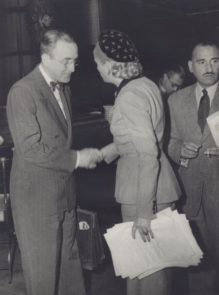 Arthur J. Altmeyer, President of the Permanent Inter-American Committee on Social Security, greeting Eva Peron, President of the Inter-American Conference on Social Security meeting in Buenos Aires. An unidentified man stands on the right, holding a cigarette.