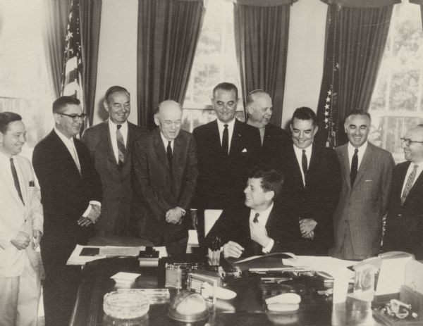 John F. Kennedy, seated, surrounded by a number of men, including Vice-President Lyndon Baines Johnson, center, standing, and Wilbur Cohen, Assistant Secretary for Legislation of Health, Education and Welfare, far right.