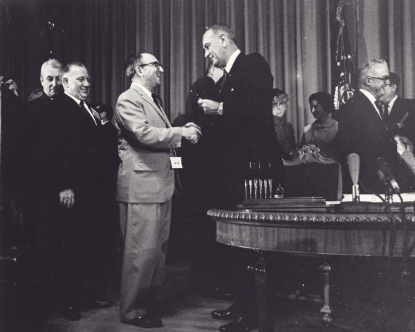 Wilbur Cohen receives a pen from President Lyndon Johnson at the formal signing of the Medicare Act part of the Social Security Amendments Act. Lady Bird Johnson is standing in the background on the right talking to another woman.