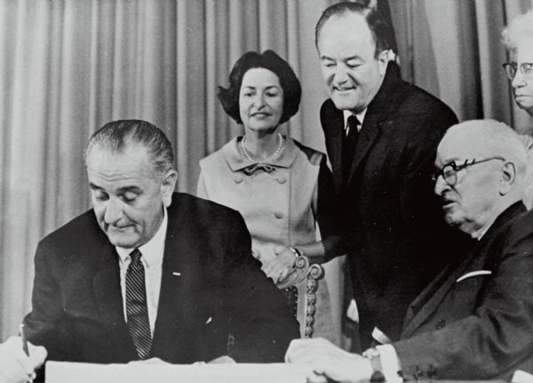 President Lyndon Baines Johnson sits at a desk signing the Medicare Act. His wife, Lady Bird Johnson, stands behind him with vice president Hubert Humphrey. There are two unidentified people on the right. This act was part of the Social Security Amendments Act of 1965.