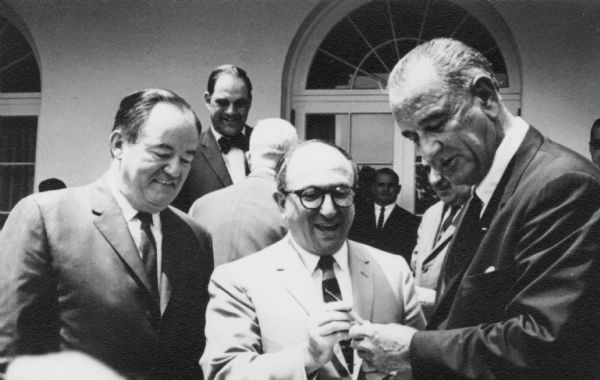 Wilbur J. Cohen receiving a pen from President Lyndon Johnson at the White House on the signing of the Older Americans Act. This act was established under the Department of Health and Human Services, where Wilbur Cohen was Under Secretary. Vice President Hubert Humphrey looks on. Congressman Scheuer of New York is in the background.