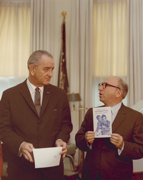 President Lyndon B. Johnson stands next to Wilbur J. Cohen who is holding up a copy of the brochure, "Social Security Information for Young Families."