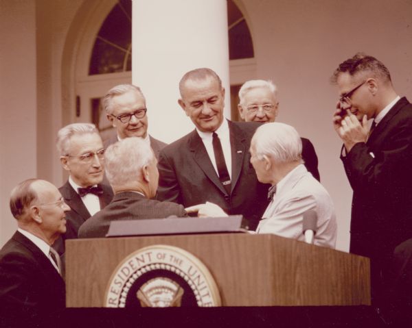 Mr. William J. Kappel of Fort Wayne, Indiana, was the 20 millionth beneficiary. President Johnson presented his first check to him at a White House ceremony. Standing on the left are Arthur Altmeyer, a former Social Security Commissioner, and Robert Ball, Commissioner of Social Security.