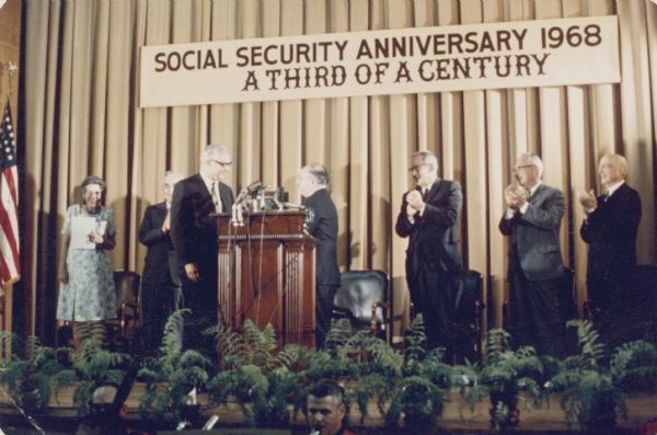 Robert Ball and Wilbur Cohen stand at the lectern during a celebration of the anniversary of Social Security. Robert Ball was Commissioner of Social Security from 1962 until 1973; Wilbur Cohen from 1935 to 1956. Both men maintained active advocacy of social security throughout the rest of their careers.