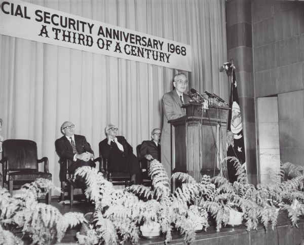 Arthur Altmeyer stands at the podium speaking about the 33rd anniversary of the Social Security bill. Wilbur Cohen and Robert Ball and an unidentified man sit behind the podium listening.