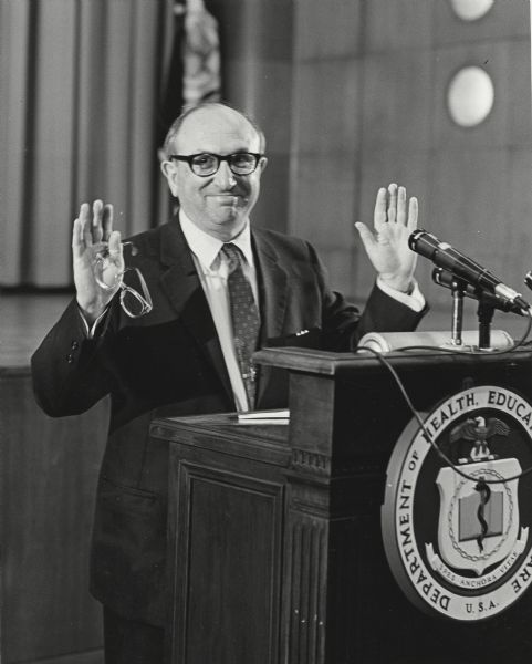 Wilbur J. Cohen, Secretary of Health, Education and Welfare, standing at a podium smiling and holding up both hands.