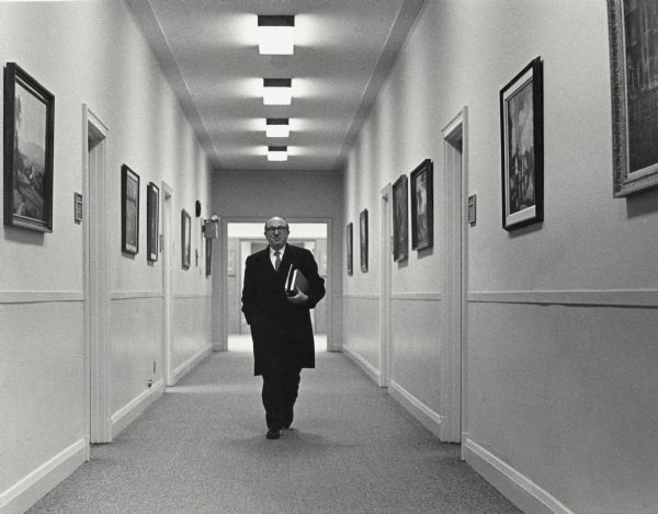 Wilbur J. Cohen, Department of Health, Education and Welfare, walks down a hallway carrying several books.