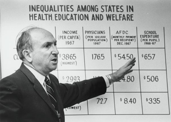 Wilbur J. Cohen, Secretary of Health, Education and Welfare, talking about inequalities while pointing at a chart. The chart has columns for Income (per capita) 1967, Physicians per 100,000 Population 1967, AFDC Monthly Payment (per recipient) Dec. 1967, and school Expediture [sic] (per pupil) 1966-67.