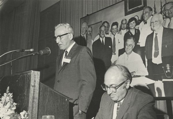 Arthur Altmeyer, standing and speaking from the podium, and Wilbur Cohen beside him, seated, at the 35th anniversary celebration of Social Security bill enactment. There is a large group photographic portrait on the wall behind Altmeyer.