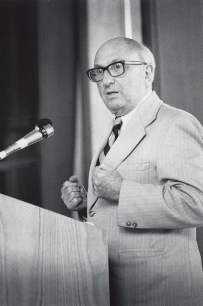 Wilbur Cohen, emphasizing a point, clenches his fists at a lectern. This is probably during a talk for Save Our Security, a non-profit coalition of national, state, and local organizations founded in 1979 by Wilbur Cohen to counter efforts to weaken the Social Security system.