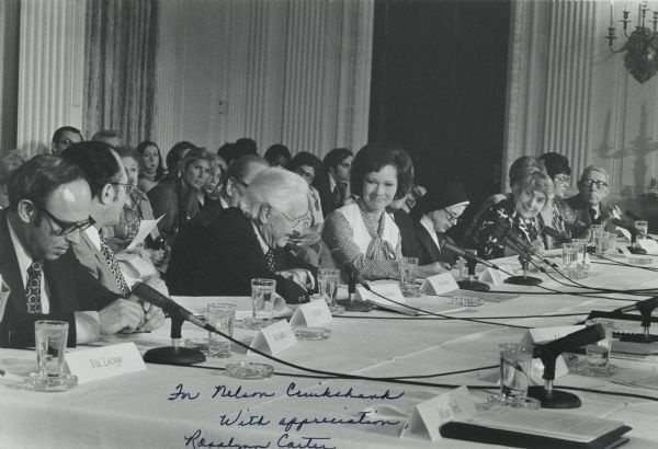 Nelson Cruikshank, leaning forward and around another person, addresses Rosalynn Carter. They are sitting at a long table with many people. On the table are microphones, name plates and water glasses. Perhaps a hearing for advancing social security benefits for people with disabilities or for benefits for the elderly.<p>Nelson Cruikshank worked for the Carter Administration from 1977 through 1980, when he left to head an education and research group with Wilbur Cohen's Save Our Security organization. As chairman of the Federal Council on Aging, he led efforts to secure and expand social security benefits for the elderly and for people with disabilities.