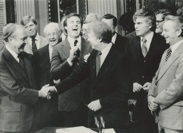 President Carter shakes hands with Senator Russell Long, D-La. after signing the Social Security bill at the White House. Laughing behind the President is Rep. Jim Guy Tucker, D-Ark.; to his left is Nelson Cruikshank. At the extreme right is Senate Majority Leader Robert Byrd, and next to him, Rep. Al Ullman, D-Ore., chairman of the House Ways and Means Committee.<p>President Jimmy Carter said:
The Social Security program is a pact between workers and their employers that they will contribute to a common fund to ensure that those who are no longer part of the work force will have a basic income on which to live. It represents our commitment as a society to the belief that workers should not live in dread that a disability, death, or old age could leave them or their families destitute." - December 20, 1977
