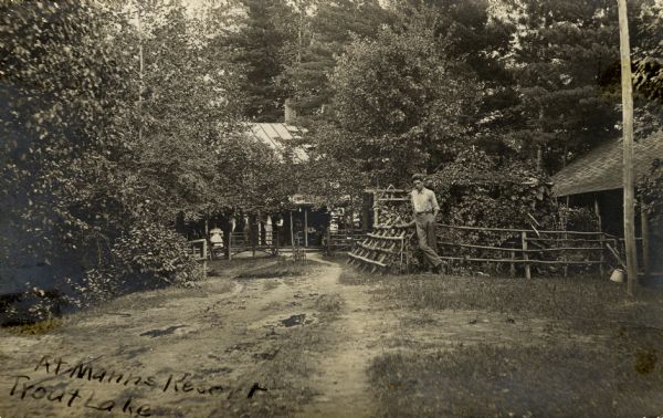 A postcard sent by C.L. (Neal) Harrington to his sister Alice Harrington in Hurley, Wisconsin. A young man is standing near a fence on the right, and behind him a group of people are sitting on a porch. Caption reads: "Mann's Resort, Trout Lake, Wis."