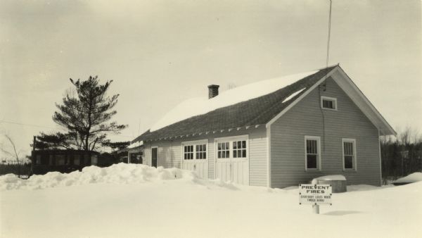 A building, surrounded in deep snow, used by Wisconsin forest rangers of district 8, perhaps in northern Wisconsin. The building includes two sets of closed garage doors. There are three windows on the right side and a window on the left side. There is also a portico on the far left side. There is a sign in front that reads: "Prevent Fires." Another building is in the background on the left.