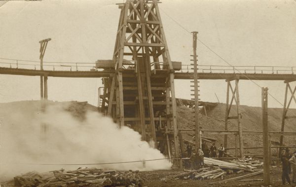 The postcard shows billowing smoke from the Hurley, Wisconsin Windsor mine shaft. Two men who were in the mine at the time lost their lives. A group of boys sit in the center foreground, and men and boys stand on the right.<p>This postcard was mailed to C.L. Harrington at Association Hall, Madison, Wisconsin.