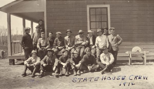 Group portrait of forest rangers outside a house. They are wearing work clothes, boots and a variety of hats. A man on the right is holding a gun and pointing it. There is a sharpening wheel in a wood frame on the right. 