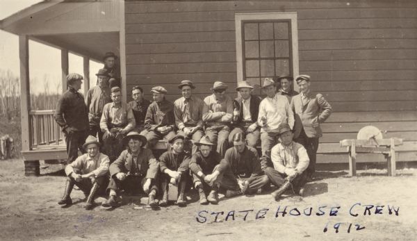 Group portrait of forest rangers outside a house. They are wearing work clothes, boots and a variety of hats. A man on the right is holding a gun and pointing it. There is a sharpening wheel in a wood frame on the right.
