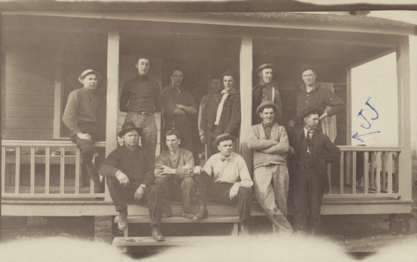 Group portrait of twelve men posing on the steps and porch of a building, probably forest rangers. The men are wearing a variety of clothing, including a bow tie, a suit, hats, shirts, pants and shoes or boots. Forest rangers planted trees and removed lumber from the Wisconsin forests. Someone has written "J.J." in blue ink, and an arrow pointing to one of the men.