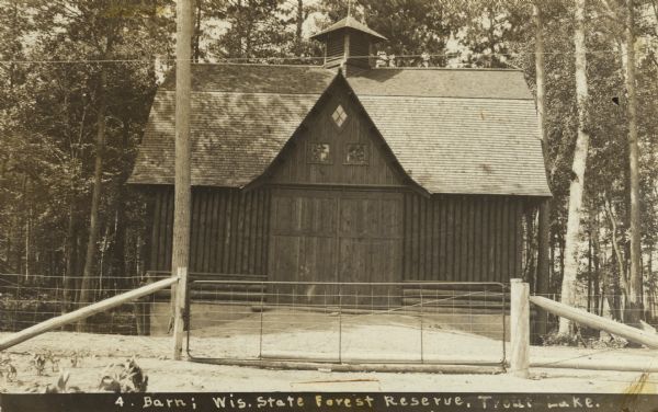 View of front of vertical log barn. Probably built in the late 19th century. It has a louvered cupola or ventilator, T-crossed barn doors and three glass windows in the upper level.<p>This barn was probably used to store forest machinery. There is a wide, strong gated entrance in the foreground. Caption reads: "Barn: Wisconsin State Forest Reserve, Trout Lake."