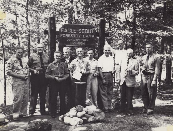 Ten men are standing in a row in front of a sign marking the 25th anniversary of the Eagle Scout Forestry Camp. They are each holding an Eagle Scout trophy. C.L. (Neal) Harrington is the fourth man from the right.<p>The Eagle Scout Forestry Camp was held in Lost Canoe Lake near Boulder Junction, Wisconsin.