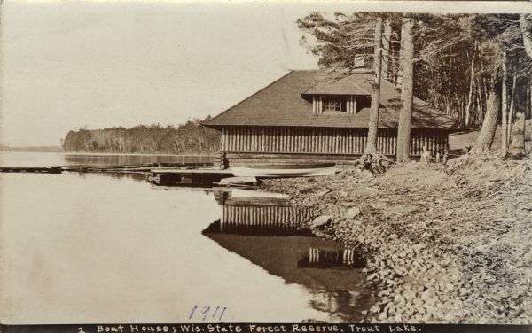 View along shoreline towards the new boat house constructed of Norway pine logs at Trout Lake. It is part of the forest ranger complex. Caption reads: "Boat House: Wisconsin State Forest Reserve, Trout Lake."