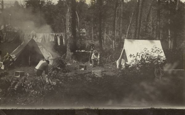 View of a campsite with two tents. One man is kneeling near a campfire blowing smoke over a tent and a line of clothes. Two other men are sitting nearby.<p>The postcard is addressed to C.L. (Neal) Harrington's father, Mr. L.L. Harrington in Hurley, Wisconsin. The handwritten text reads:<p>"Dear Pop:-  I'm dropping you this card mid-weekly for as you probably noticed I'm short on coin. I ordered the new suit the other day and will be called on to pay for it soon. Will you send me the usual amount. I am feeling fine. Hope you are all well. Neal."
