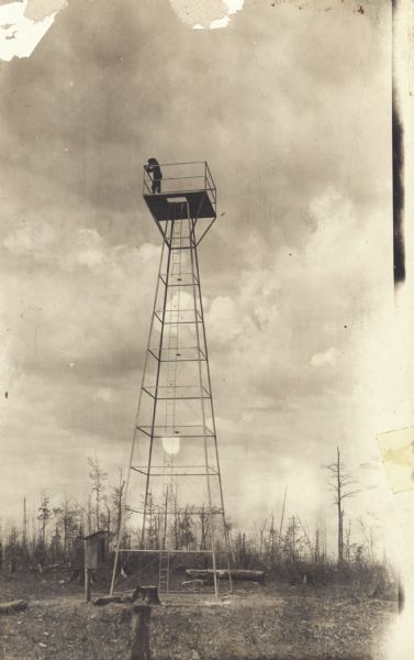 View towards tower, with a man standing on the platform on the top. This postcard has a handwritten notation on the back: "One of the first forest protection lookouts erected — a windmill tower with a platform on top."